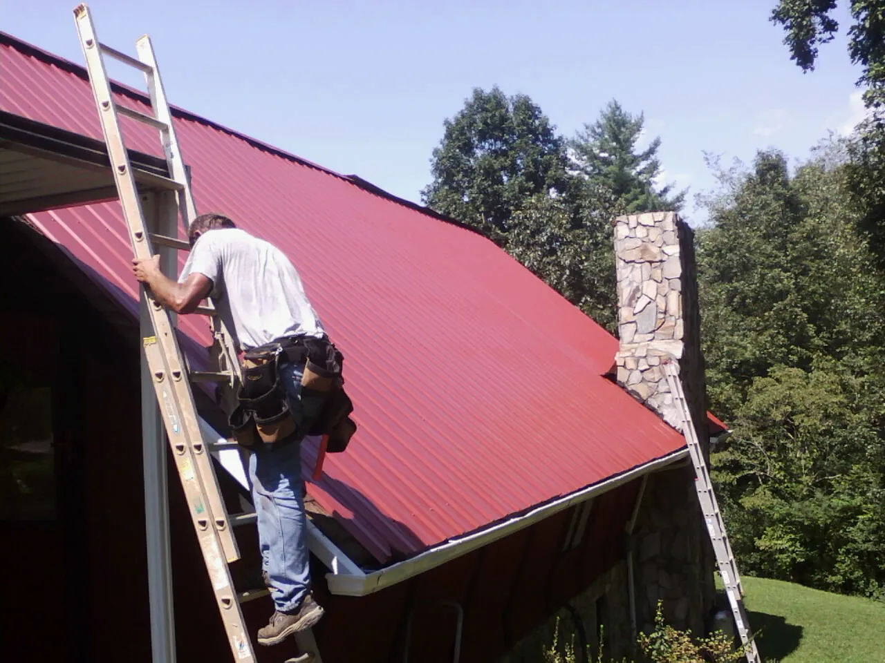 Roofer on a ladder beside a red metal roof and stone chimney — Soffit & Fascia Repair by Roofing Brothers in Saint Cloud